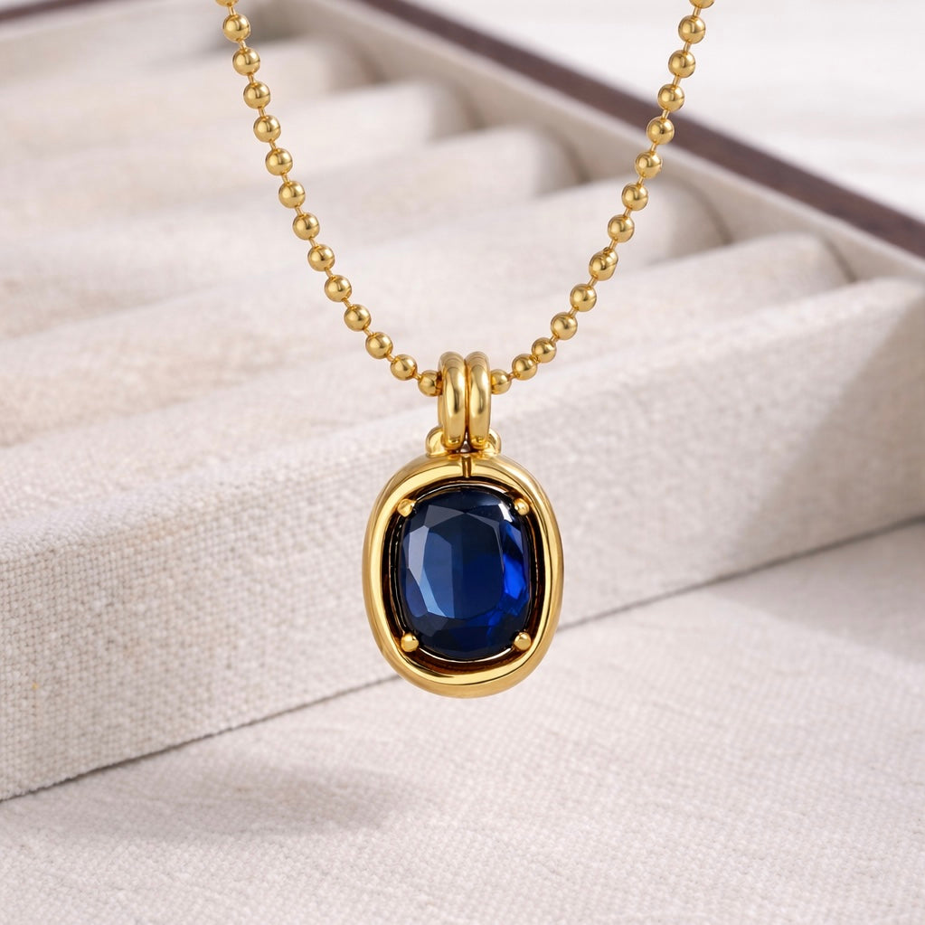 Close-up of Noelle gold beaded chain necklace with deep blue gemstone pendant on jewelry display cushion