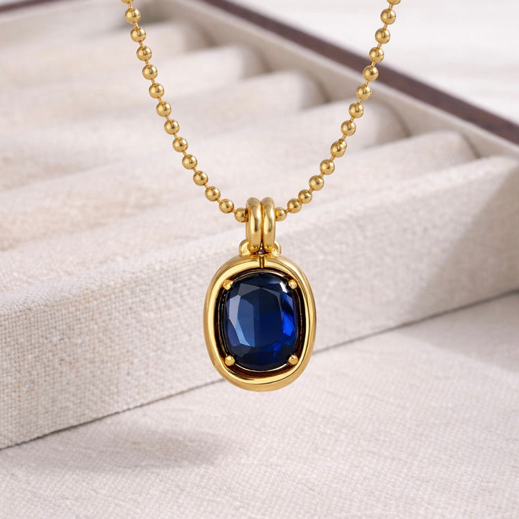 Close-up of Noelle gold beaded chain necklace with deep blue gemstone pendant on jewelry display cushion